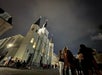 A group of people gather in front of an illuminated historic church with tall spires under a cloudy night sky.