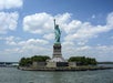 The Statue of Liberty on Liberty Island, surrounded by trees, stands against a partly cloudy sky, with water in the foreground.