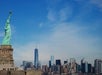 The Statue of Liberty stands in the foreground with a clear view of the New York City skyline under a blue sky.