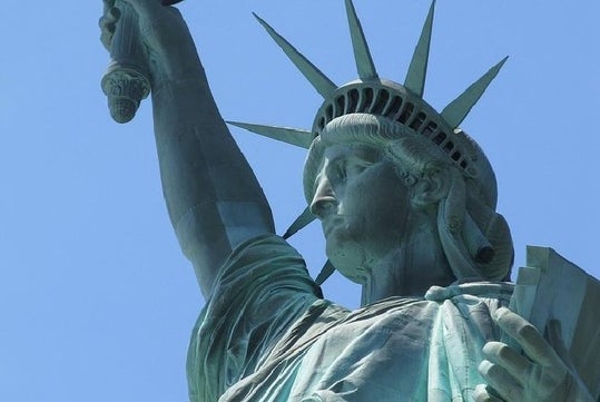 Close-up view of the Statue of Liberty's face and upper torso, with her arm raised holding a torch, against a clear blue sky.
