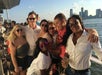A group of people posing together on a boat, with a city skyline and river in the background. They are smiling and some are holding drinks.