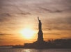Silhouette of the Statue of Liberty against a sunset sky, with water in the foreground and trees in the background.
