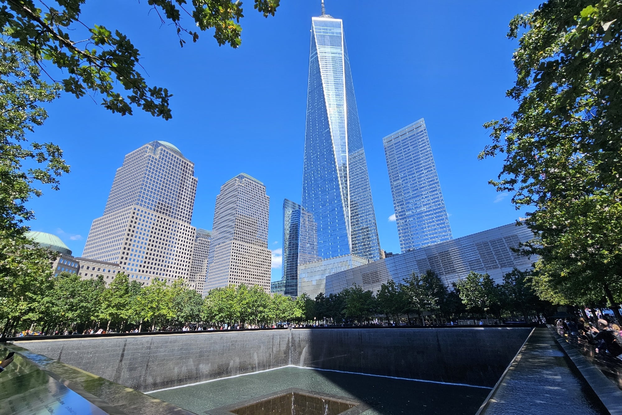 One World Trade Center and surrounding buildings overlook the 9/11 Memorial reflecting pool under a clear blue sky, framed by green trees.