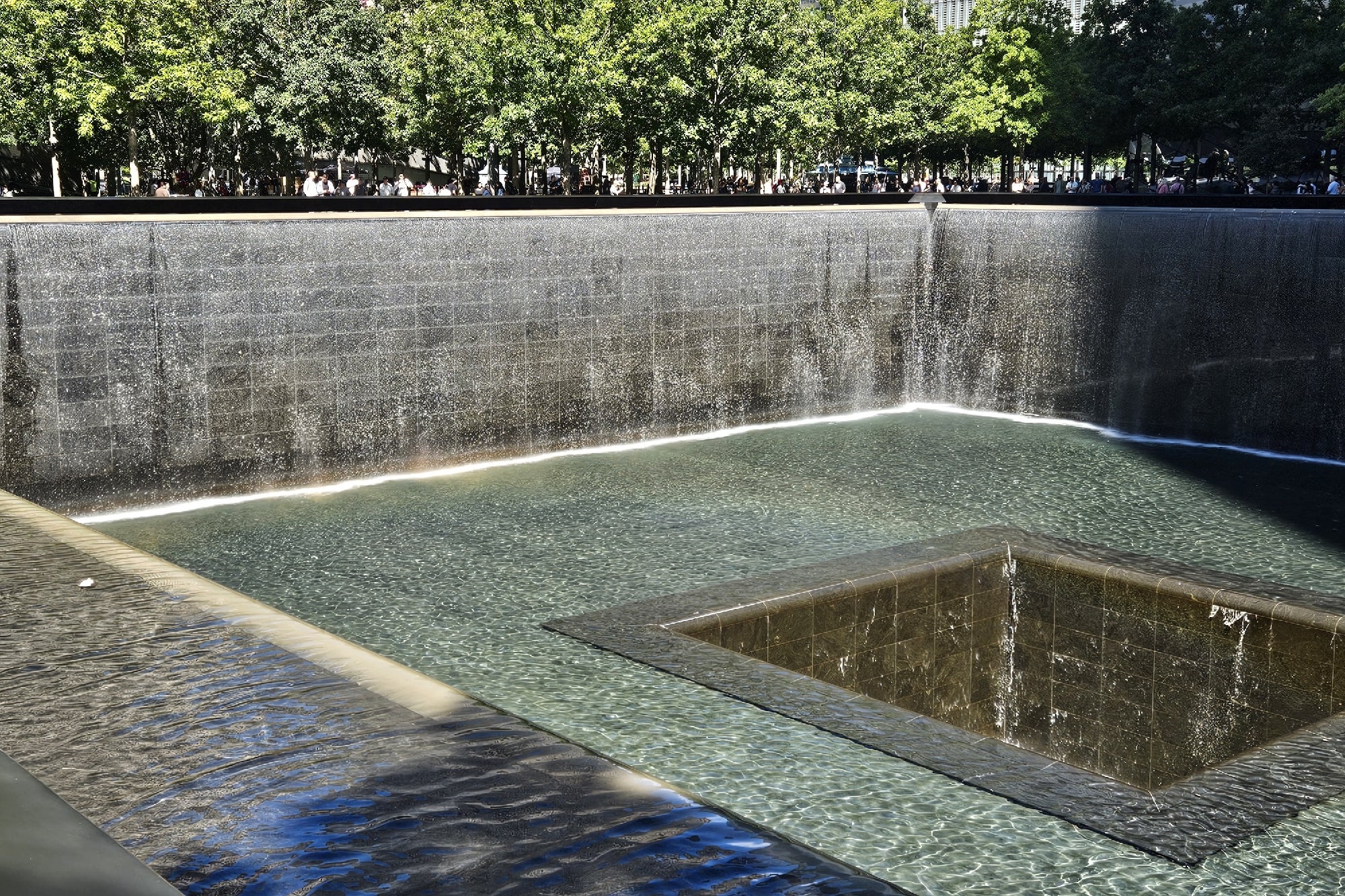 A large square memorial fountain with water flowing down black stone walls, trees in the background, and people gathered along the edge.