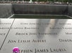 Names inscribed on the edge of a memorial fountain at the 9/11 Memorial in New York City, with a purple and white flower placed by one name.