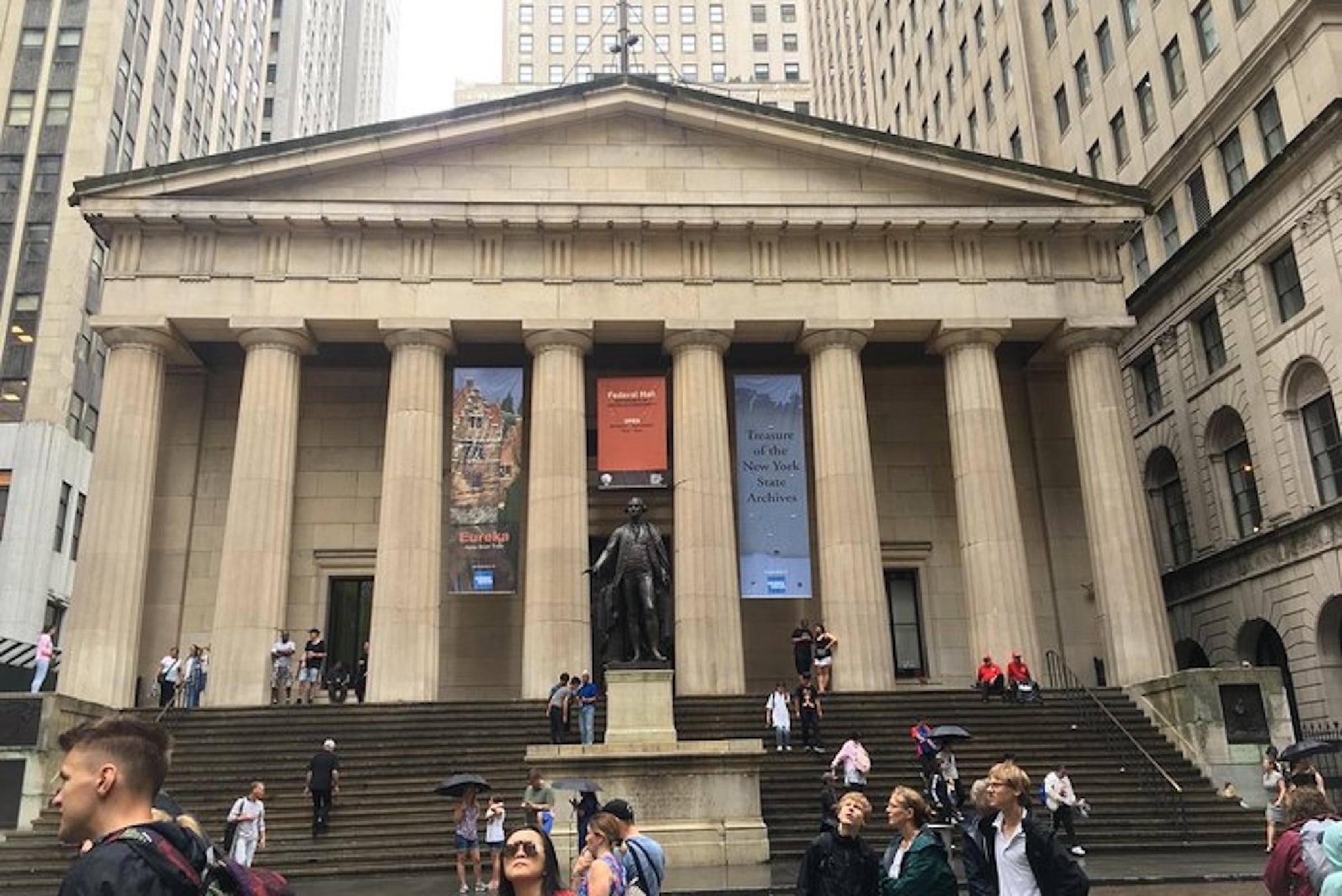 A neoclassical building with tall columns and a statue in front, surrounded by people; banners hang beside the entrance.
