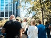 A group of people stand outdoors near trees and tall glass buildings on a sunny day.