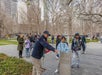 A tour guide gestures towards a plaque titled "Survivor Tree" as a group of people listen and take photos in an outdoor urban park with buildings and leafless trees in the background.