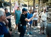 A group of people stands outdoors in a park, listening to a person in a black shirt and cap who is speaking and gesturing with one hand.