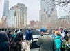 A group of people stands around the 9/11 Memorial reflecting pool in New York City on an overcast day, with tall buildings in the background.