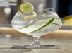 Elegant cocktail glass filled with clear liquid, garnished with a lime wedge and cucumber slices, on a coaster.
