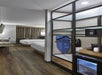 Hotel room with a wooden floor, two beds, TV, desk, ironing board, and a small fridge. Shelving unit holds a safe and tray with a coffee maker. Curtain covers a window in the background.