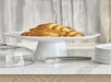Plate with croissants on a pedestal stand, next to a stack of white plates and metal tongs on a marble countertop.