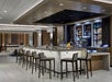 A modern bar with high stools surrounds the counter. Behind the bar, shelves display various bottles. The space features wooden accents and soft lighting.