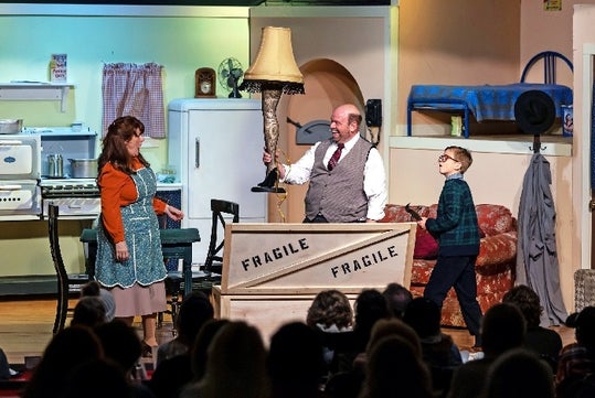A father standing behind a box holding the iconic leg lamp with the mother to his left and to his right on stage at the A Christmas Story Dinner Show.