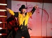 A man in a yellow and black cowboy shirt and hat performs on stage, pointing outward with both hands, with a guitarist playing in the background.
