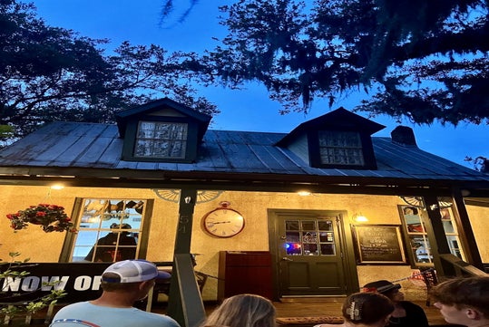 People gather outside a lit, rustic house with a porch at dusk. A large clock hangs near the door, and a sign indicates the place is now open. Trees frame the scene.