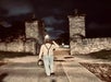 A person wearing suspenders and a hat walks at night through a stone gate, carrying a bag, with streetlights and trees visible in the background.