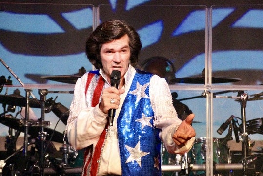 A man wearing a sequined, red, white, and blue vest with stars sings into a microphone onstage in front of a drum set.