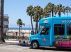 A bright blue shuttle bus with "Hollywood" on its side is parked near a beach. Palm trees and a lifeguard tower are visible in the background under a clear blue sky.