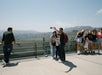 A group of people takes selfies and poses on an observation deck with a hilly landscape in the background on a clear day.