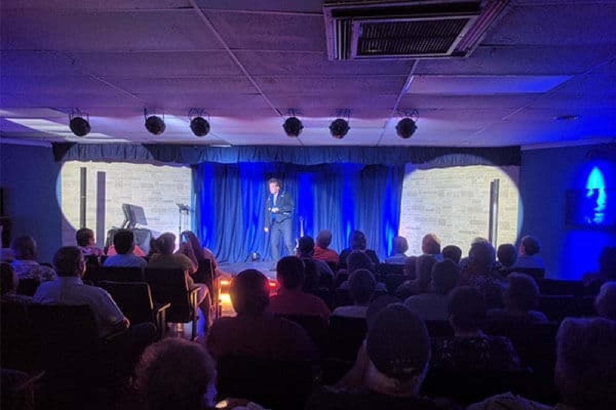 An audience watches a performer on a small stage with blue curtains and spotlighting.