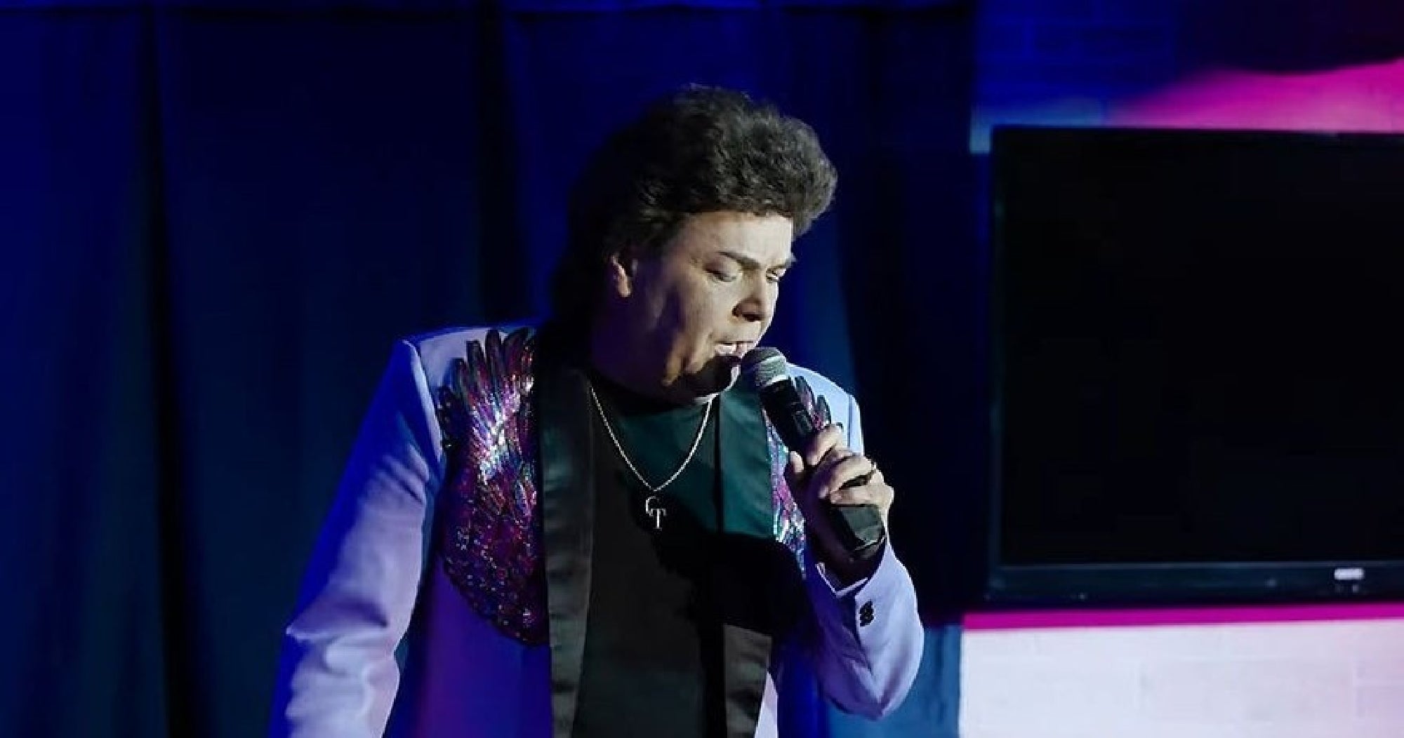Person singing into a microphone on stage, wearing a jacket with sequin details.