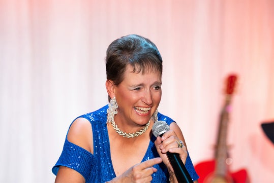 Person in a blue sequin dress is holding a microphone, smiling and pointing. A guitar is visible in the background.