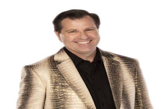 Man smiling, wearing a gold, reptile-patterned blazer over a black shirt on a white background.