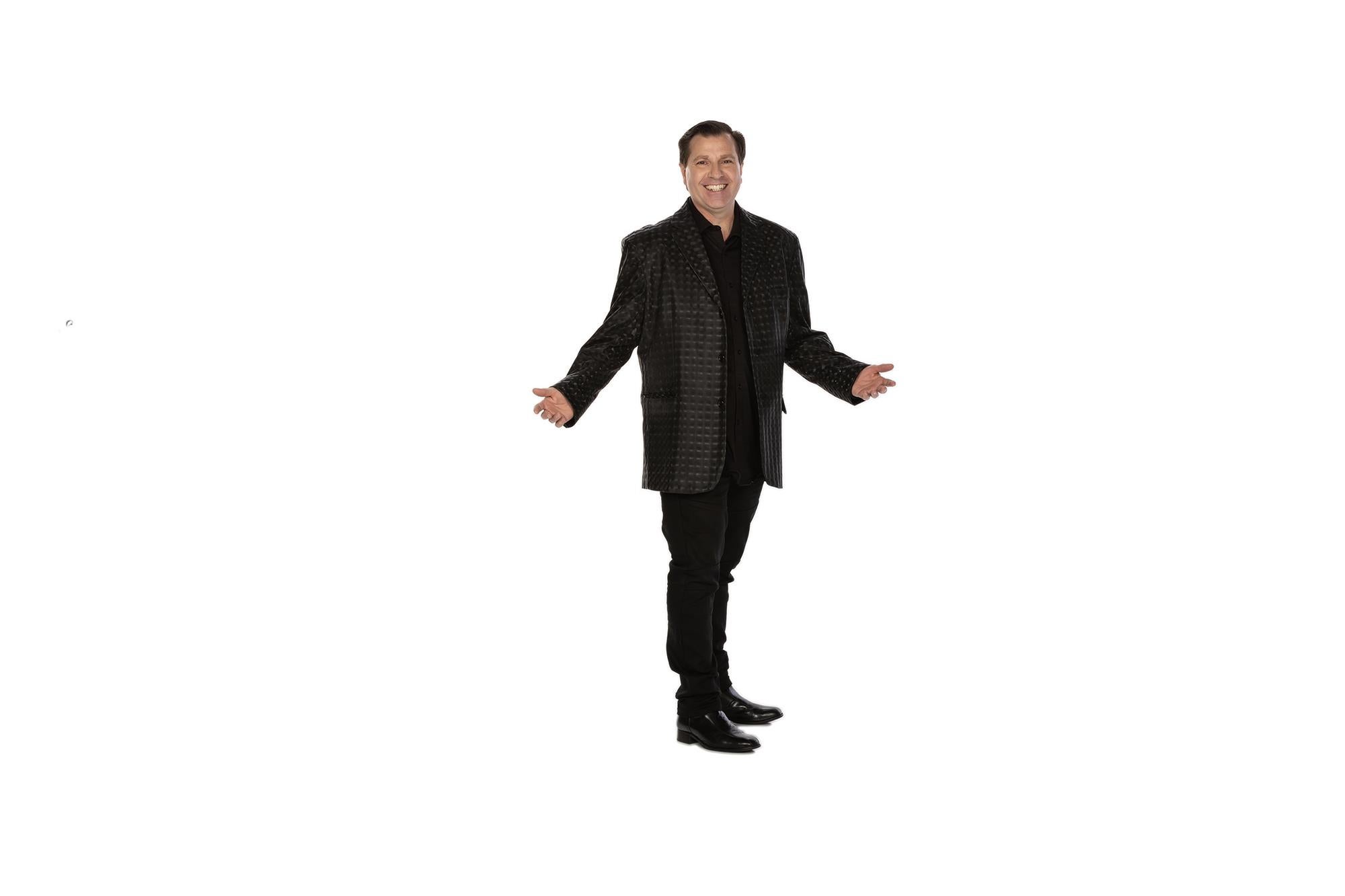 Man in a dark patterned jacket and black pants smiling with arms open against a white background.