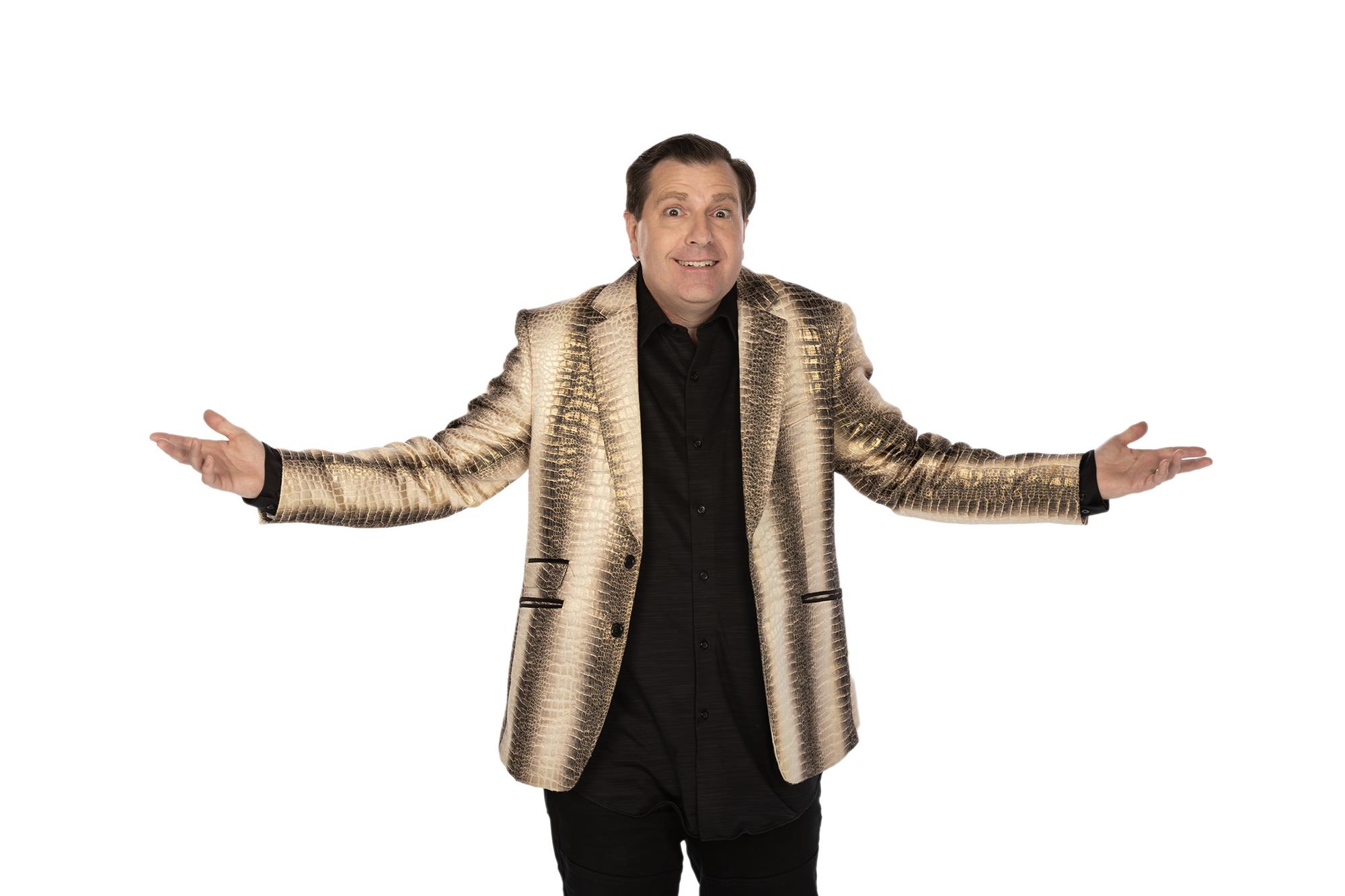 Man in a gold jacket with a textured design, wearing black pants and shirt, stands with arms outstretched against a white background.