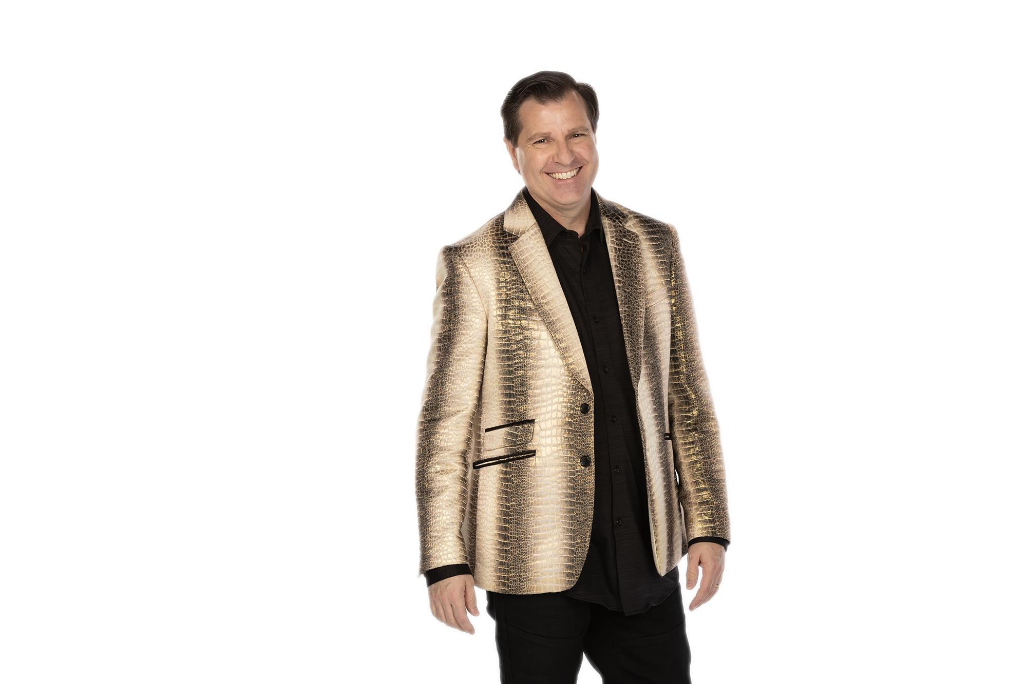 A man in a patterned gold jacket and black shirt stands smiling against a plain white background.
