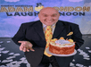Adam London in a black suit posing on stage with his congratulatory 6th year anniversary cake featuring rubber ducks on top.