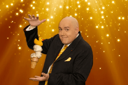 Adam London in a black suit maneuvering an ice cream with the rubber duck on top that seems to be levitating between his hands.
