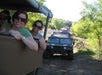 People in an open vehicle on a safari trail are smiling, with another off-road vehicle in the background surrounded by greenery.