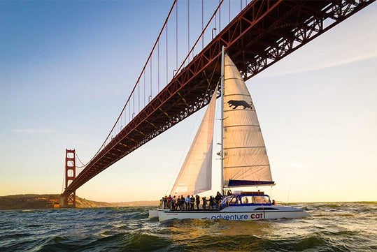 A sailboat with people onboard passes under the Golden Gate Bridge in San Francisco during daylight.