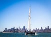 A sailboat moves across the water with a city skyline in the background under a clear blue sky.