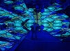 A person stands in a mirrored room filled with blue and green kaleidoscopic light patterns, creating multiple reflections of people and the geometric designs.