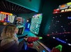 A woman plays a shooting arcade game while other people play Pac-Man and Connect 4 in a dimly lit arcade.