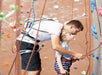 An adult helps a young child fasten a harness at an indoor climbing gym, with colorful climbing holds and ropes visible in the background.