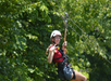 Person wearing a helmet and harness ziplining through a lush green forest, smiling and making a peace sign with fingers.