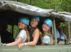 Three young women wearing helmets smile at the camera while seated in an open-top vehicle, surrounded by trees in a forested area.
