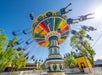 Colorful swing ride with people seated, spinning under a clear blue sky. Trees and a yellow building are in the background.