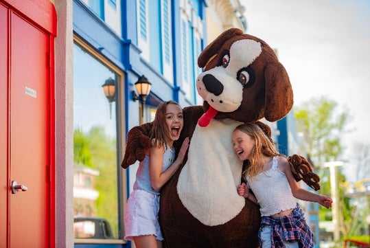 Two young girls laughing and hugging the Bernie Bernard mascot with colorful buildings on the background at Adventureland.