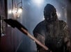 A menacing figure in dirty overalls and a grotesque mask wields a bloodied weapon in a dimly lit, foggy hallway.