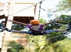 Person wearing a helmet and harness ziplining, with arms and legs spread, surrounded by trees and a wooden structure in the background.