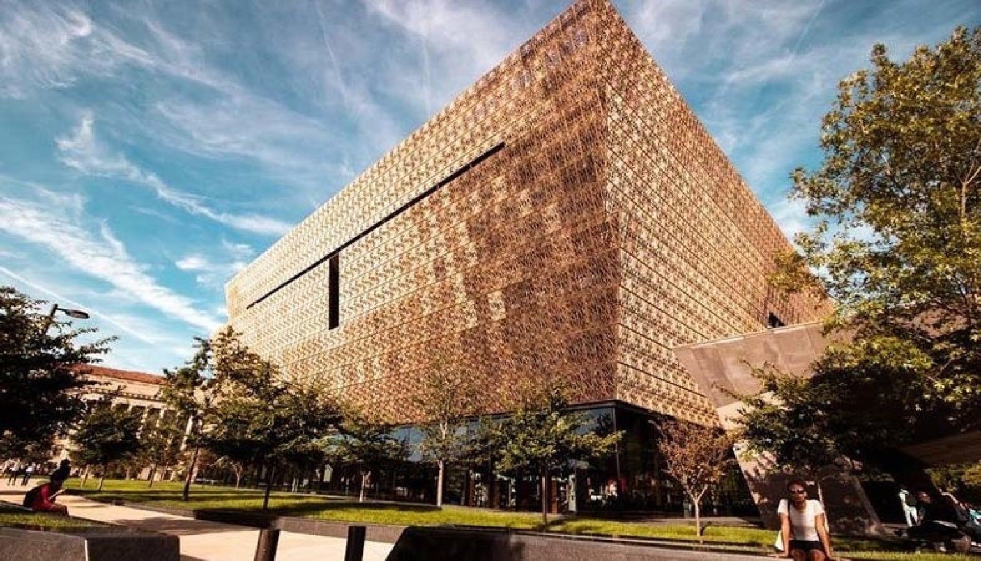 A large contemporary building with a patterned bronze facade, surrounded by trees and people walking, under a partly cloudy sky.
