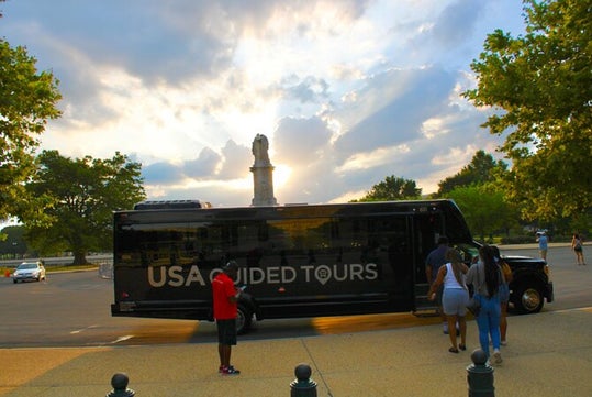 A USA Guided Tours bus is parked on a street with trees and a monument in the background. People are walking nearby under a sky with sun rays breaking through clouds.