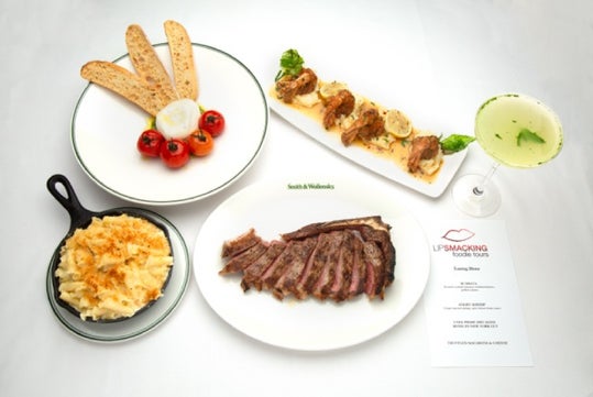 A plated steak, macaroni and cheese in a skillet, crostini appetizers, roasted tomatoes with bread, a cocktail, and a printed menu are arranged on a white surface.