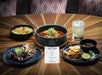 A wooden table set with four dishes, a margarita cocktail, and a printed menu. The dishes include grilled meat, tacos, rice, and a stew, with garnishes on each plate.